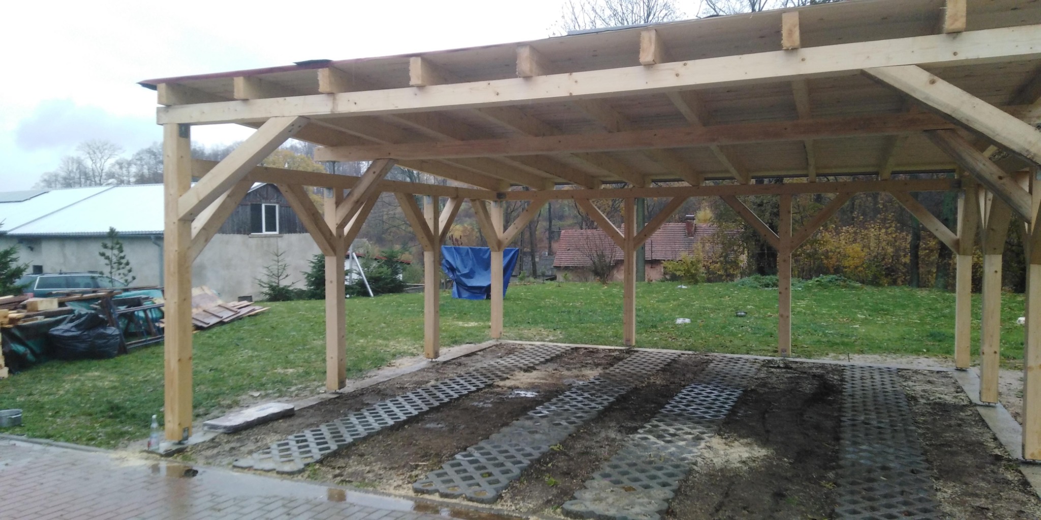 Surowa drewniana konstrukcja wiaty samochodowej z widoczną więźbą dachową, niezadaszona, z podłożem z ażurowych płyt betonowych, w tle budynek mieszkalny i drzewa.