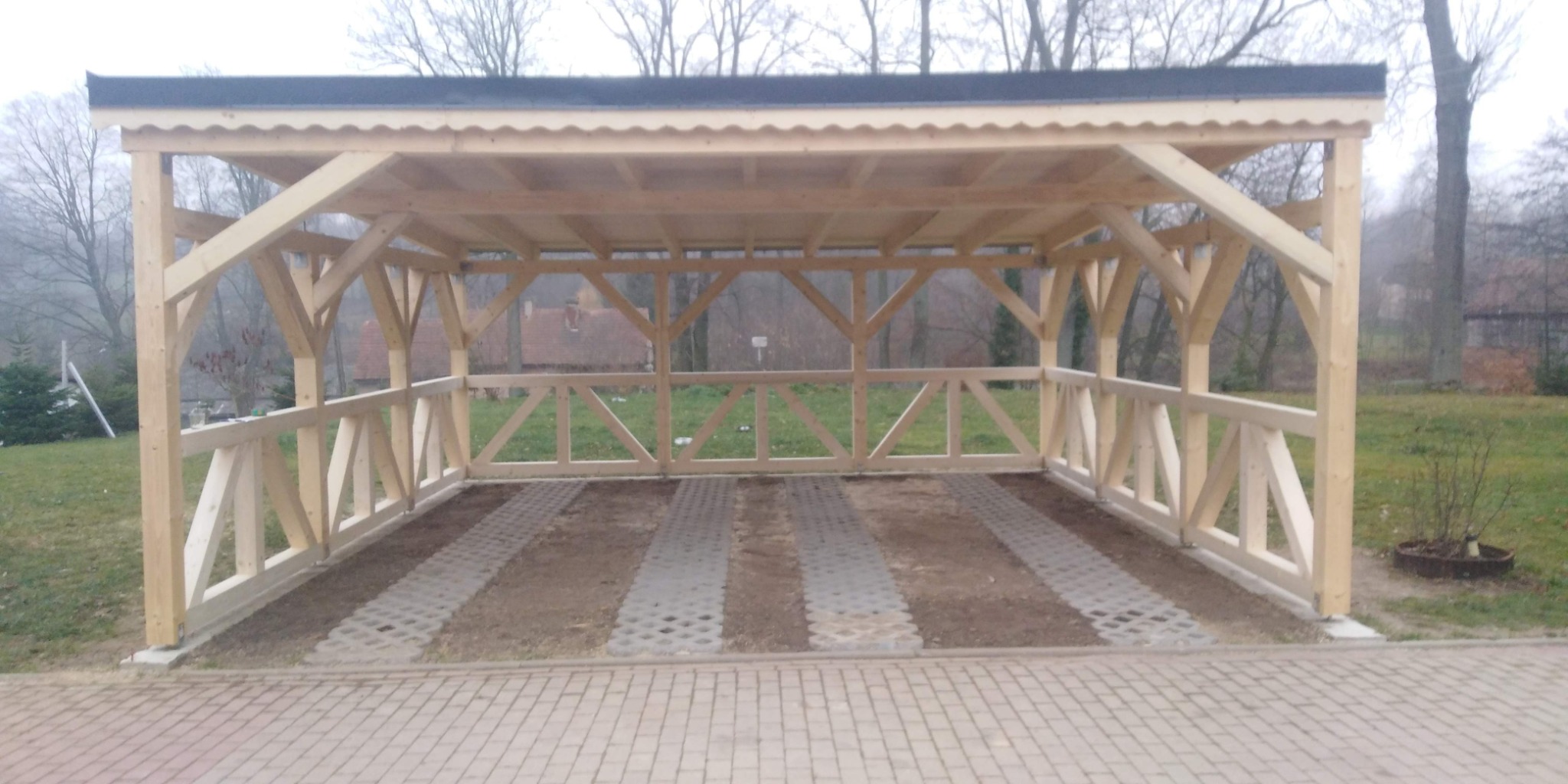 Drewniana wiata garażowa z czarnym dachem, wykończona drewnianą balustradą, widok z zewnątrz na kostkę brukową i trawnik w pochmurny dzień.