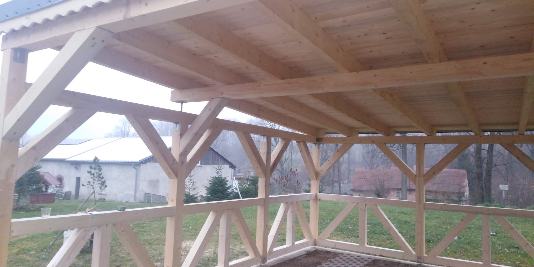 Surowa drewniana konstrukcja zadaszenia tarasu z widoczną więźbą dachową i balustradą, w tle zabudowania gospodarcze.