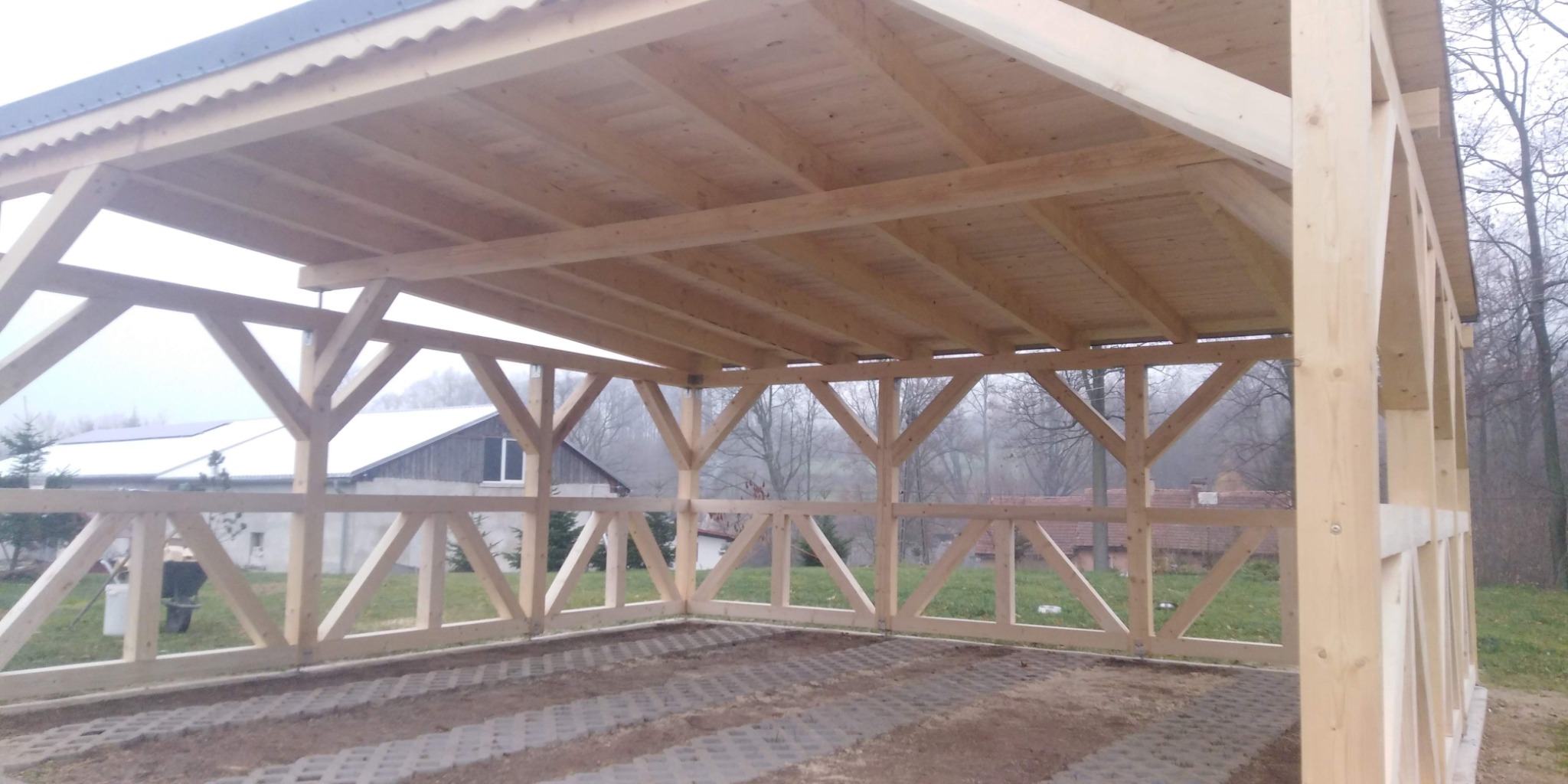 Drewniana konstrukcja zadaszenia w trakcie budowy, widoczne elementy więźby dachowej, słupy nośne i dekoracyjne obramowanie, w tle budynki mieszkalne i drzewa.