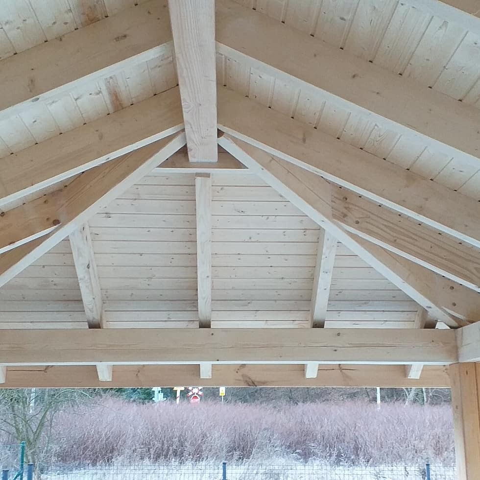 Widok od dołu na jasną, drewnianą konstrukcję dachu z widoczną więźbą i deskowaniem, w tle widoczny krajobraz z drzewami bez liści.
