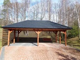 Drewniana wiata garażowa z ciemnym dachem krytym dachówką bitumiczną, z ażurowymi ścianami, na posesji z kostki brukowej, na tle drzew bezlistnych.