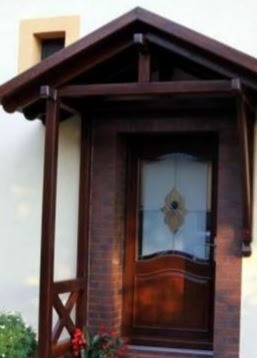 Zadaszenie nad wejściem do domu, wykonane z ciemnego drewna, wsparte na słupach z ozdobnymi krzyżakami, mur z cegły klinkierowej do wysokości drzwi, jasna elewacja budynku.