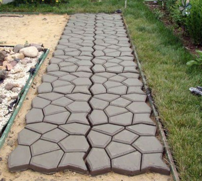 Świeżo ułożona ścieżka z betonowych płyt o nieregularnych, geometrycznych kształtach, na piaskowym podłożu, oddzielona metalowym obrzeżem od trawnika. Płyty jeszcze mokre, w trakcie schnięcia.