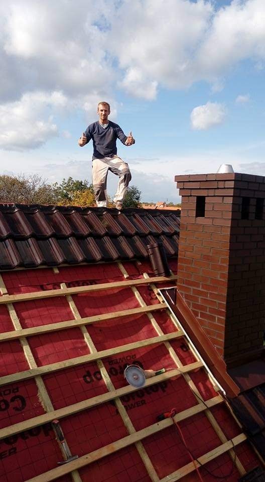 Mężczyzna na dachu z nową dachówką, kominem z cegły i widoczną konstrukcją więźby, z narzędziami na membranie dachowej, pozujący z uniesionymi kciukami na tle błękitnego nieba z chmurami.