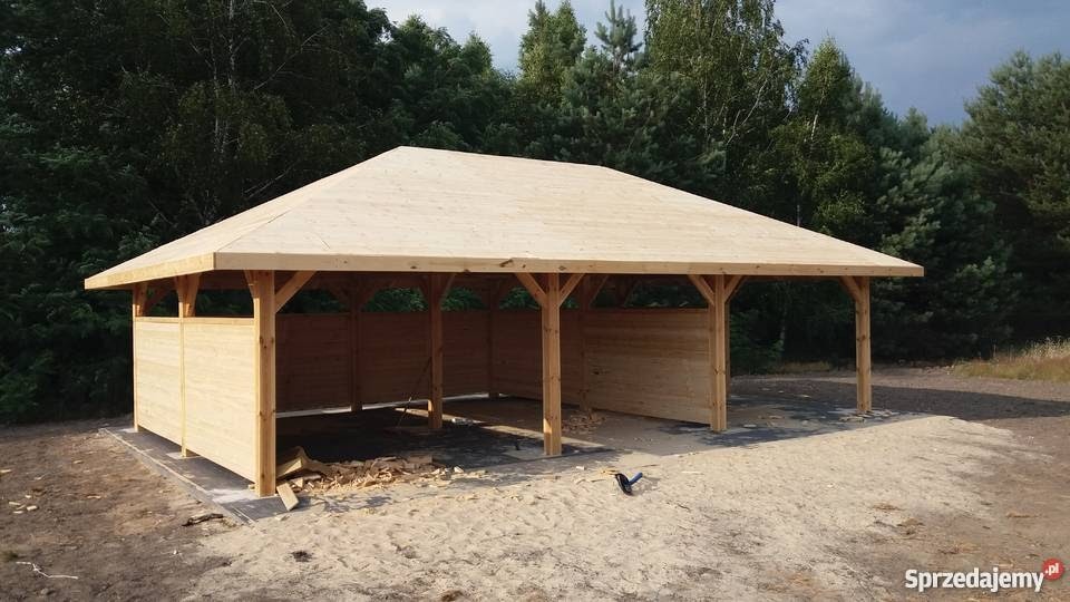 Surowa drewniana altana z płaskim dachem, częściowo obudowana deskami, widoczna na tle zieleni, z drobnymi odpadami po obróbce drewna wokół.