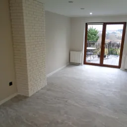 Pusty pok&oacute;j po remoncie z szarą podłogą imitującą marmur, białą kolumną z cegły i wyjściem na balkon z widokiem na wzg&oacute;rza.
