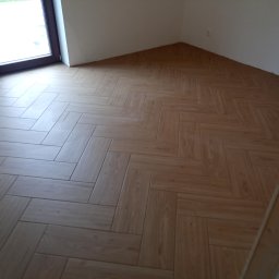 Welus - Ułożona w jodełkę podłoga z płytek imitujących drewno, w jasnym odcieniu, widok z góry na nową posadzkę w pomieszczeniu z oknem.