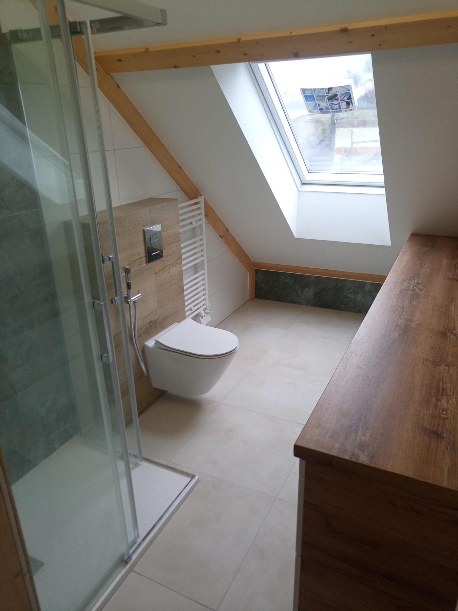 Nowoczesna łazienka na poddaszu z kabiną prysznicową, podwieszaną toaletą, grzejnikiem drabinkowym i drewnianą szafką pod umywalkę, oświetlona oknem dachowym.