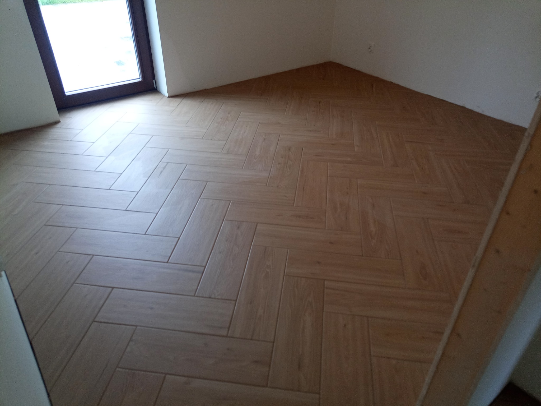 Ułożona w jodełkę podłoga z płytek imitujących drewno, w jasnym odcieniu, widok z góry na nową posadzkę w pomieszczeniu z oknem.