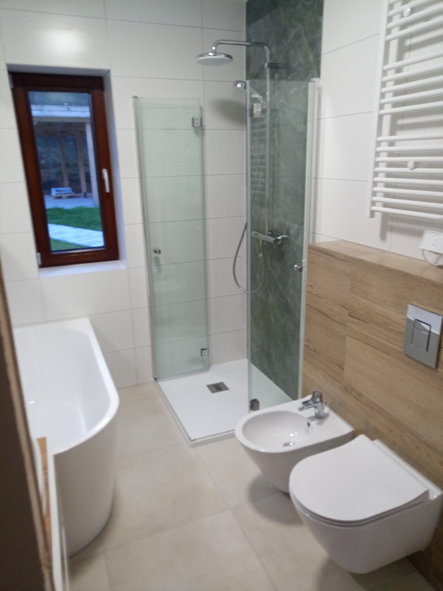 Nowoczesna łazienka z kabiną prysznicową z zieloną ścianką, białą wanną, bidetem i toaletą podwieszaną, okno z widokiem na ogród.