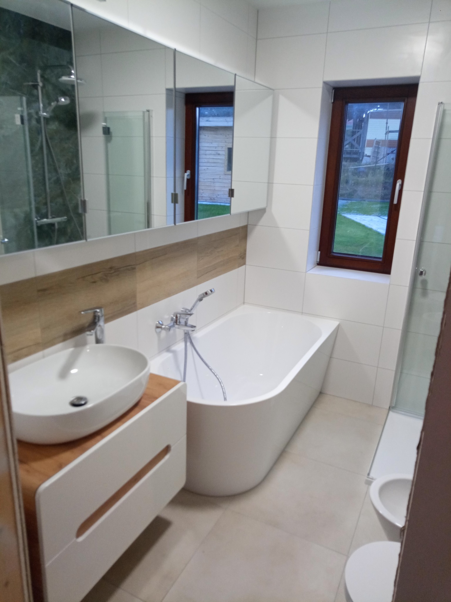 Nowoczesna łazienka z białymi płytkami, drewnopodobnym akcentem nad umywalką, owalną wanną i oknem z widokiem na zewnątrz.