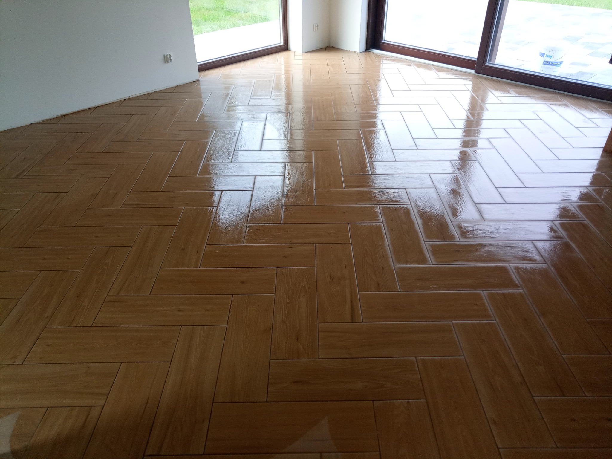 Błyszcząca, ułożona w jodełkę podłoga z płytek imitujących drewno, widok z niskiego kąta, duże okna w tle.