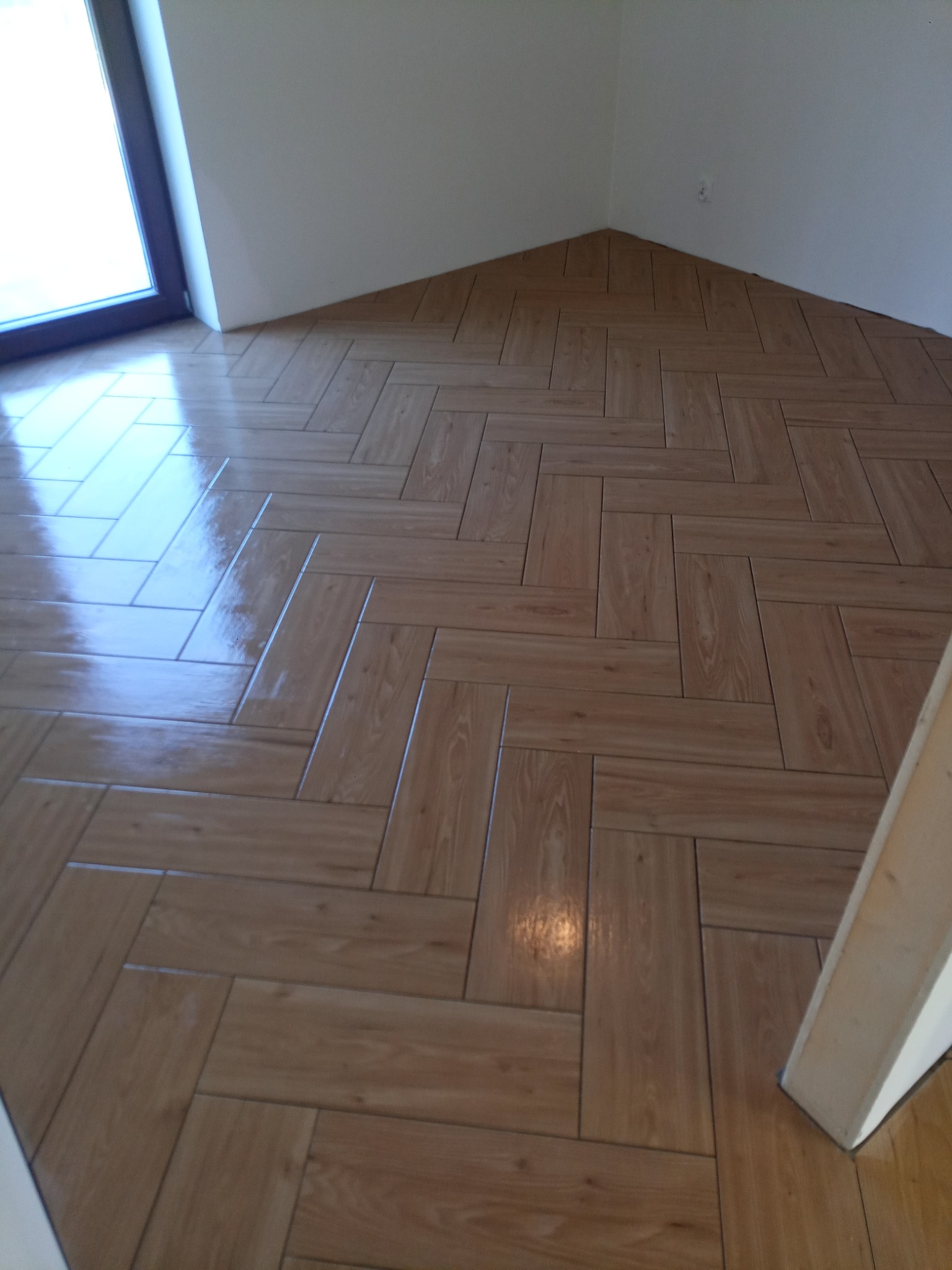 Ułożona w jodełkę, błyszcząca podłoga z płytek imitujących drewno, widok z góry, fragment okna i ściany w tle.