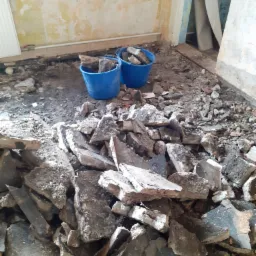 Stos gruzu i fragmentów betonu na podłodze w trakcie remontu, dwa niebieskie wiadra częściowo wypełnione gruzem w tle, ściany z widocznymi śladami starej farby.