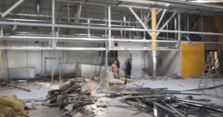 Wnętrze hali w trakcie demontażu, widoczne elementy konstrukcyjne, gruz i dwóch pracowników w oddali.