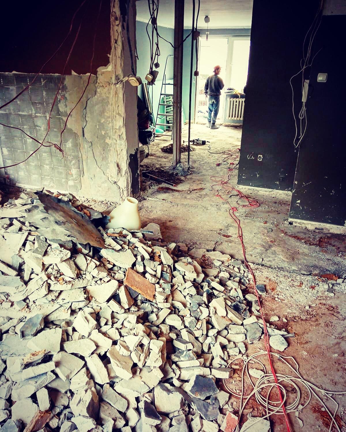 Sterta gruzu z fragmentami płytek i cegieł w pomieszczeniu w trakcie demontażu, widoczne przewody elektryczne i sylwetka osoby w oddali.