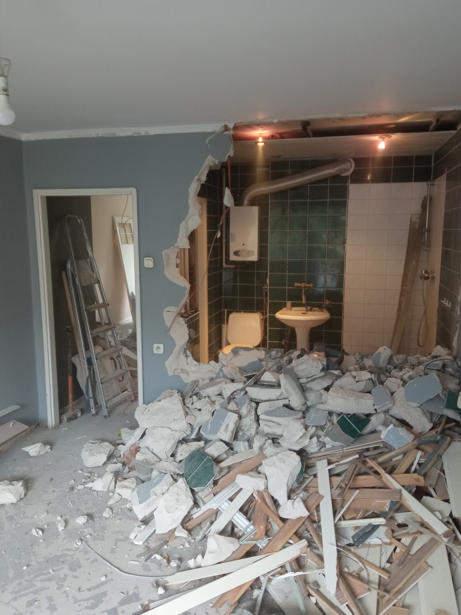 Fragment ściany między pokojem a łazienką po wyburzeniu, widoczne pozostałości gruzu, płytek i instalacji, drabina oparta o ścianę w pokoju obok.