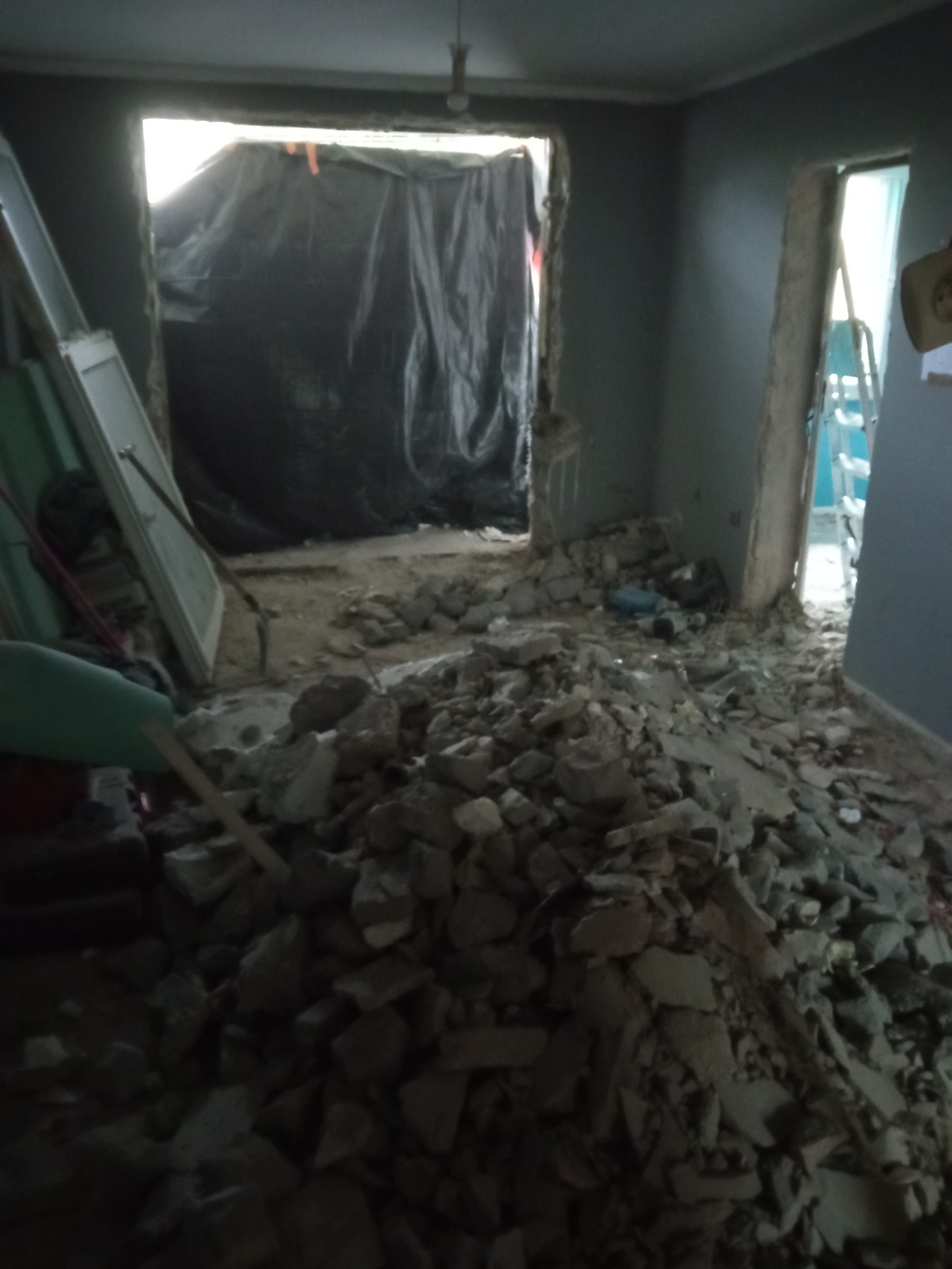 Pokój po wyburzeniu ściany działowej, z gruzem na podłodze i zabezpieczeniem folią w przejściu, widoczna drabina oparta o ścianę.