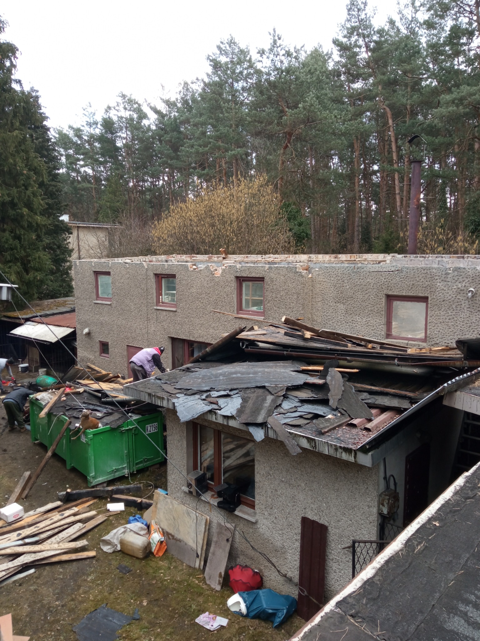 Demontaż dachu i fragmentu ściany budynku z fakturą baranka, widoczny kontener na odpady i pracownicy w trakcie pracy, otoczenie leśne w tle.