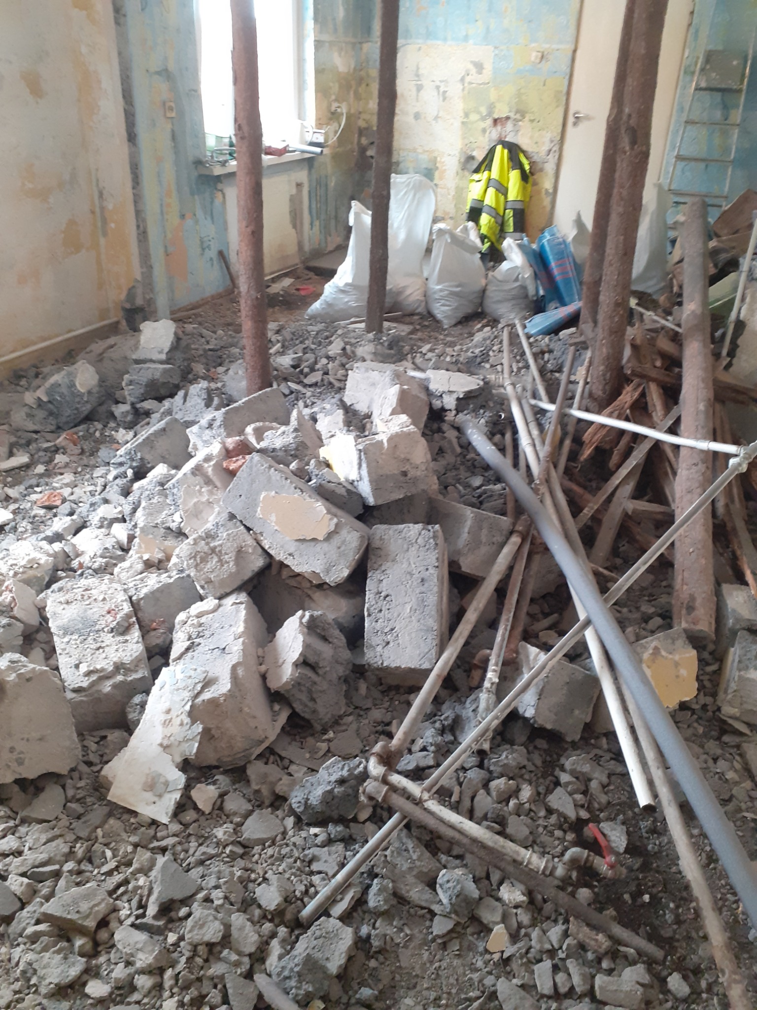 Wnętrze w Zawierciu po demontażu z gruzem, fragmentami cegieł, rurami i elementami drewnianymi na podłodze, widoczne podtrzymujące drewniane stemple i worki z odpadami w tle.