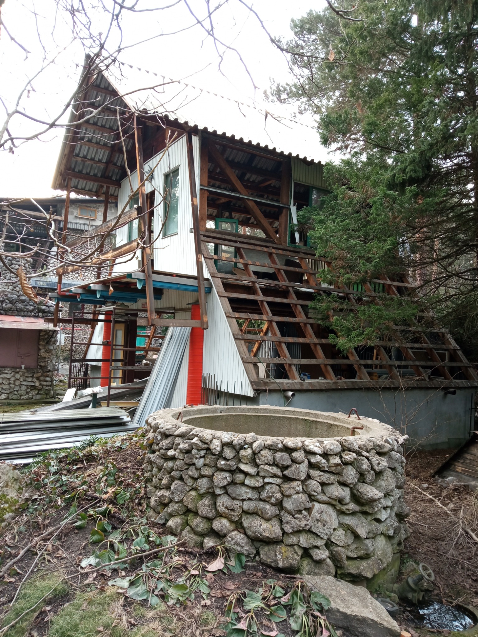 Niedokończony budynek z odsłoniętą konstrukcją dachu, widoczne elementy drewniane i białe panele, w tle drzewa i studnia z kamienia polnego, na pierwszym planie leżące płyty.