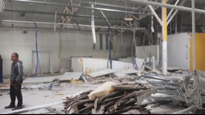 Wnętrze hali w trakcie rozbiórki, widoczne zdemontowane elementy konstrukcyjne, kable zwisające z sufitu, pracownik w tle po lewej stronie.