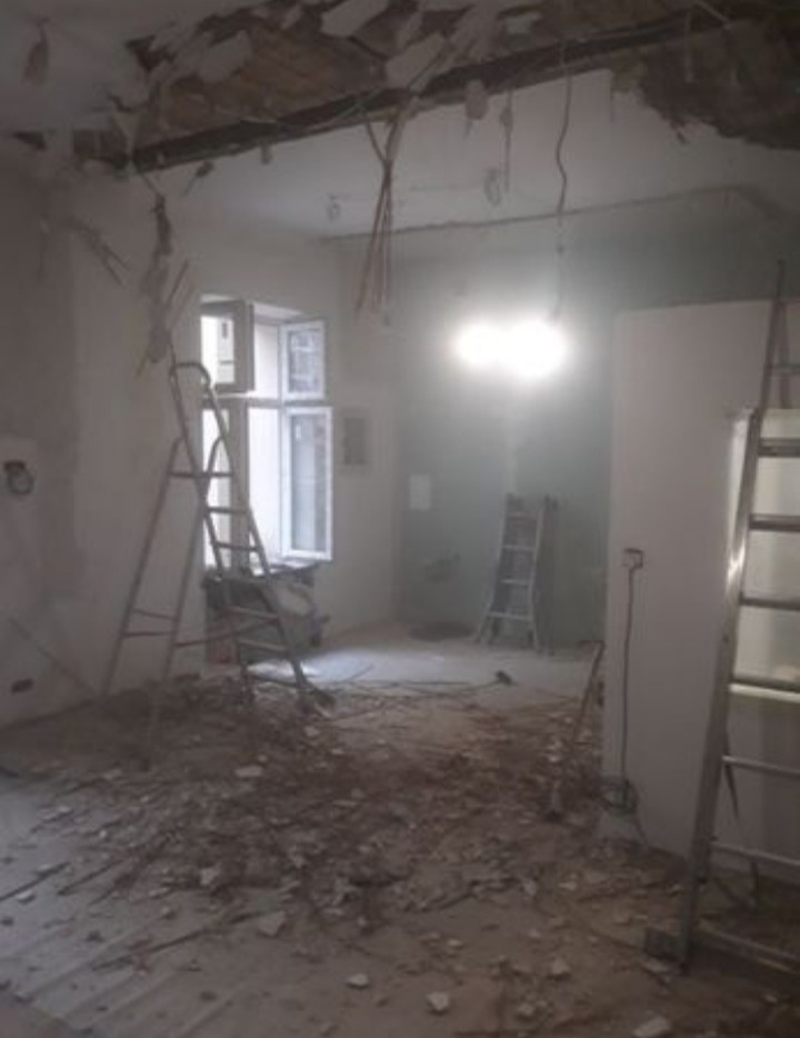 Pokój w trakcie prac rozbiórkowych: uszkodzony sufit, gruz na podłodze, drabiny opierające się o ściany i okno.