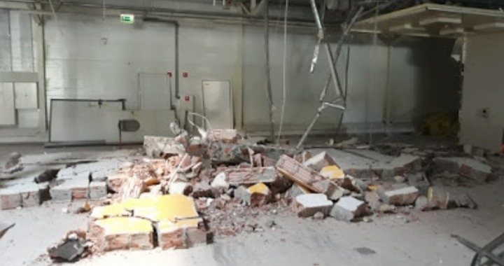 Wnętrze hali po częściowym wyburzeniu ściany, z gruzem ceglanym i elementami konstrukcyjnymi na podłodze, widoczne fragmenty instalacji i rusztowania.