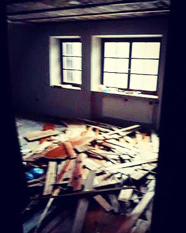 Wnętrze pokoju w trakcie demontażu, z widocznymi deskami i fragmentami konstrukcji na podłodze, dwa okna w ścianie, słabe oświetlenie.