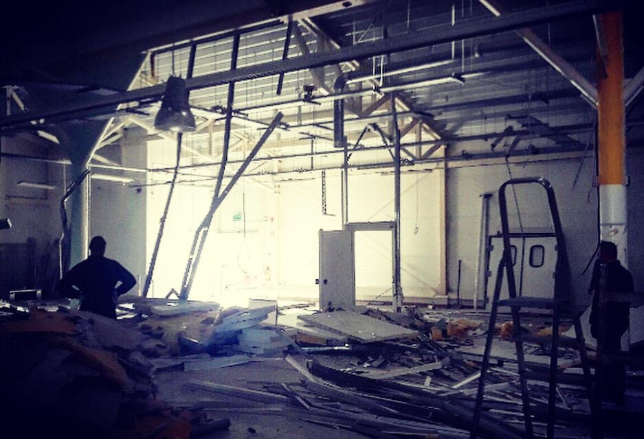 Wnętrze hali po demontażu, widoczne resztki gruzu, metalowe elementy konstrukcyjne, drabina i sylwetki osób na tle jasnego otworu.