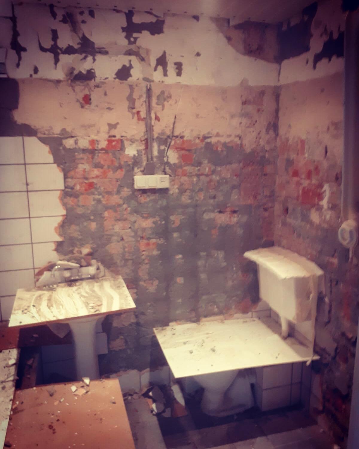 Łazienka w trakcie demontażu: częściowo usunięte płytki, odsłonięta cegła, widoczne instalacje elektryczne, zdemontowane umywalki i toaleta, zabezpieczone płytami, surowy stan pomieszczenia.