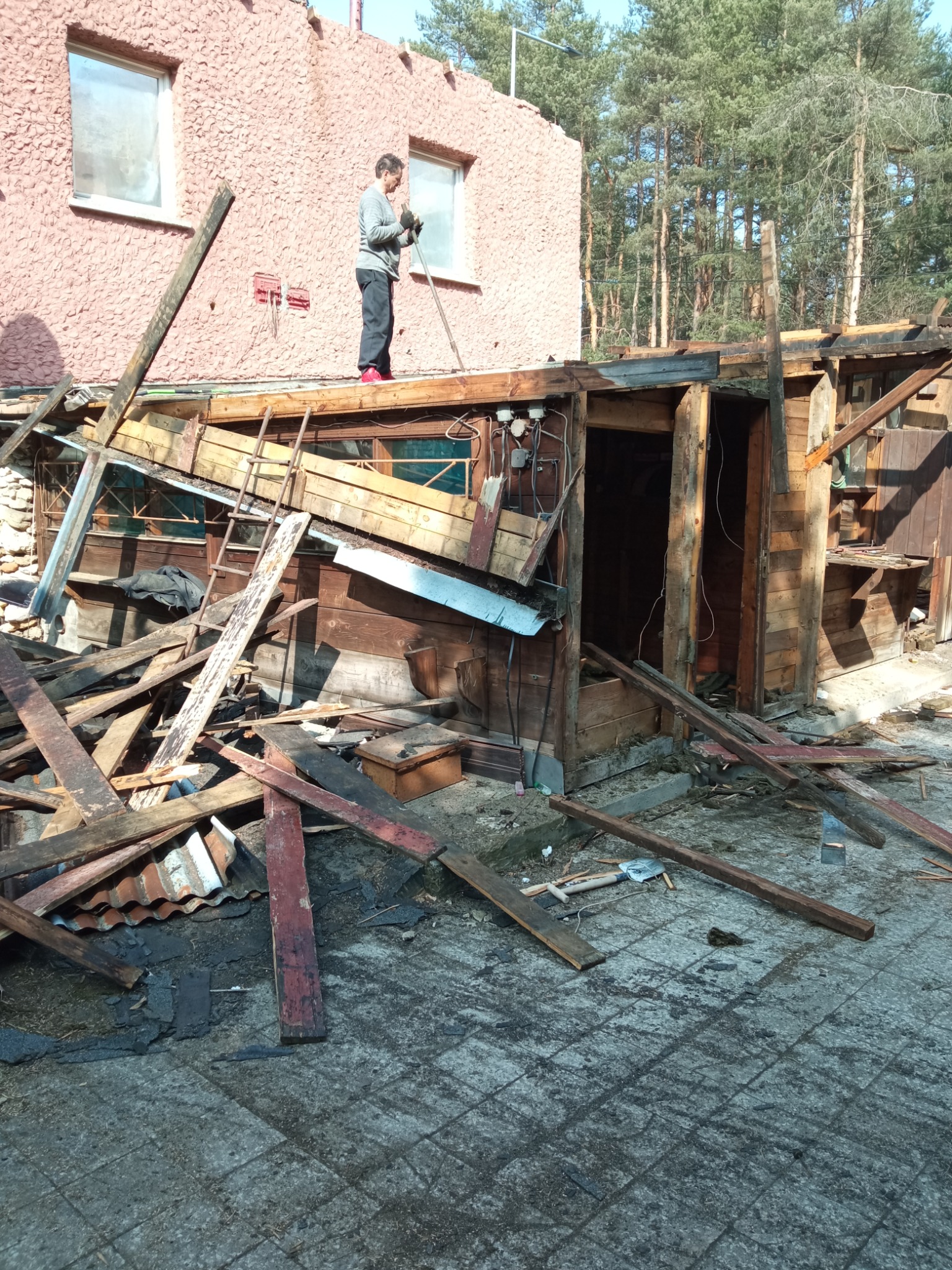 Mężczyzna w szarym swetrze i czerwonych butach stoi na dachu częściowo rozebranego budynku, trzymając narzędzie. Wokół porozrzucane deski i gruz, w tle budynek z różową elewacją i drzewa.