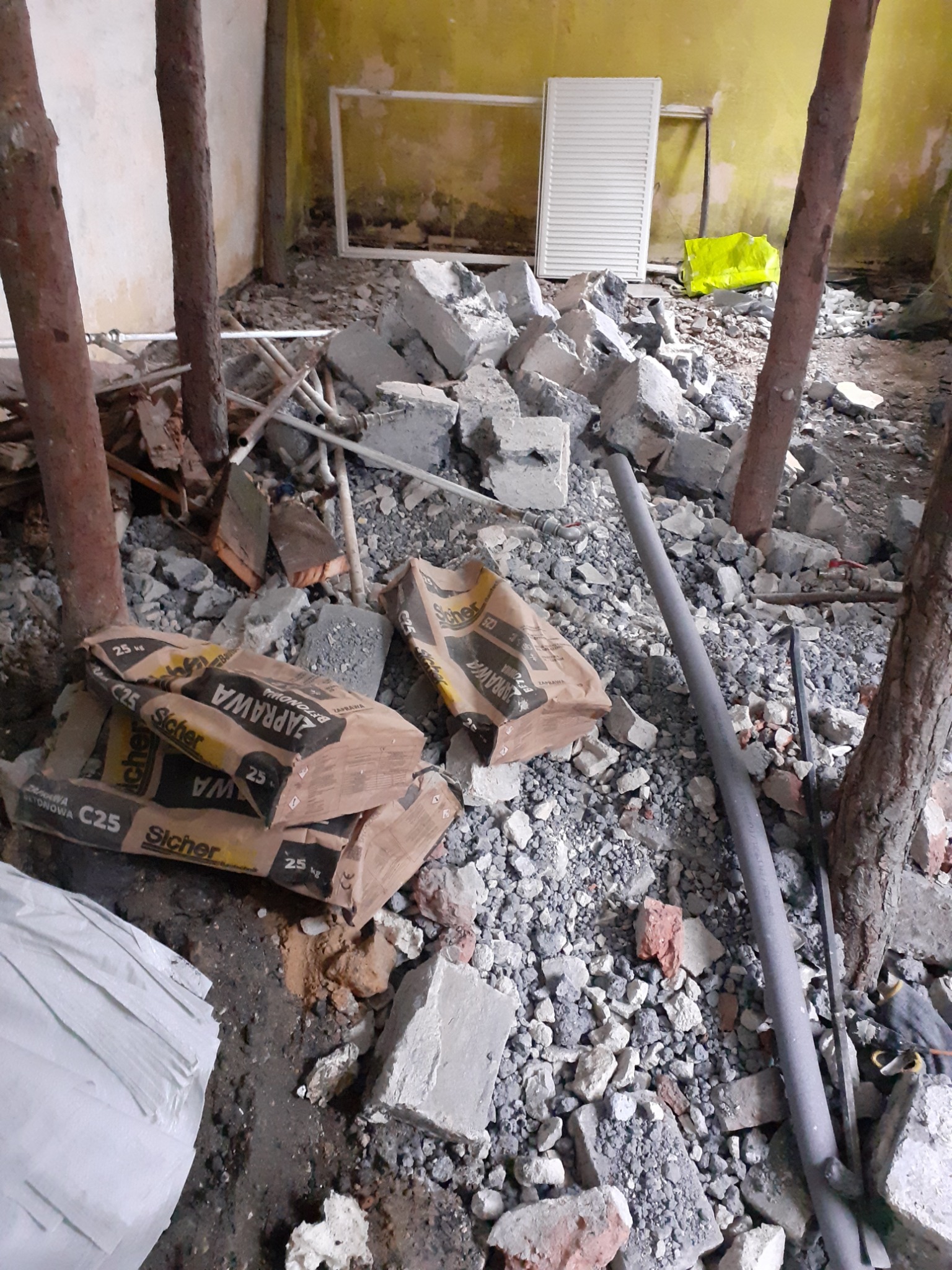 Wnętrze w trakcie rozbiórki z gruzem, workami z zaprawą murarską C25, fragmentami instalacji i drewnianymi słupami podtrzymującymi strop, widoczne resztki starej instalacji grzewczej i okno.
