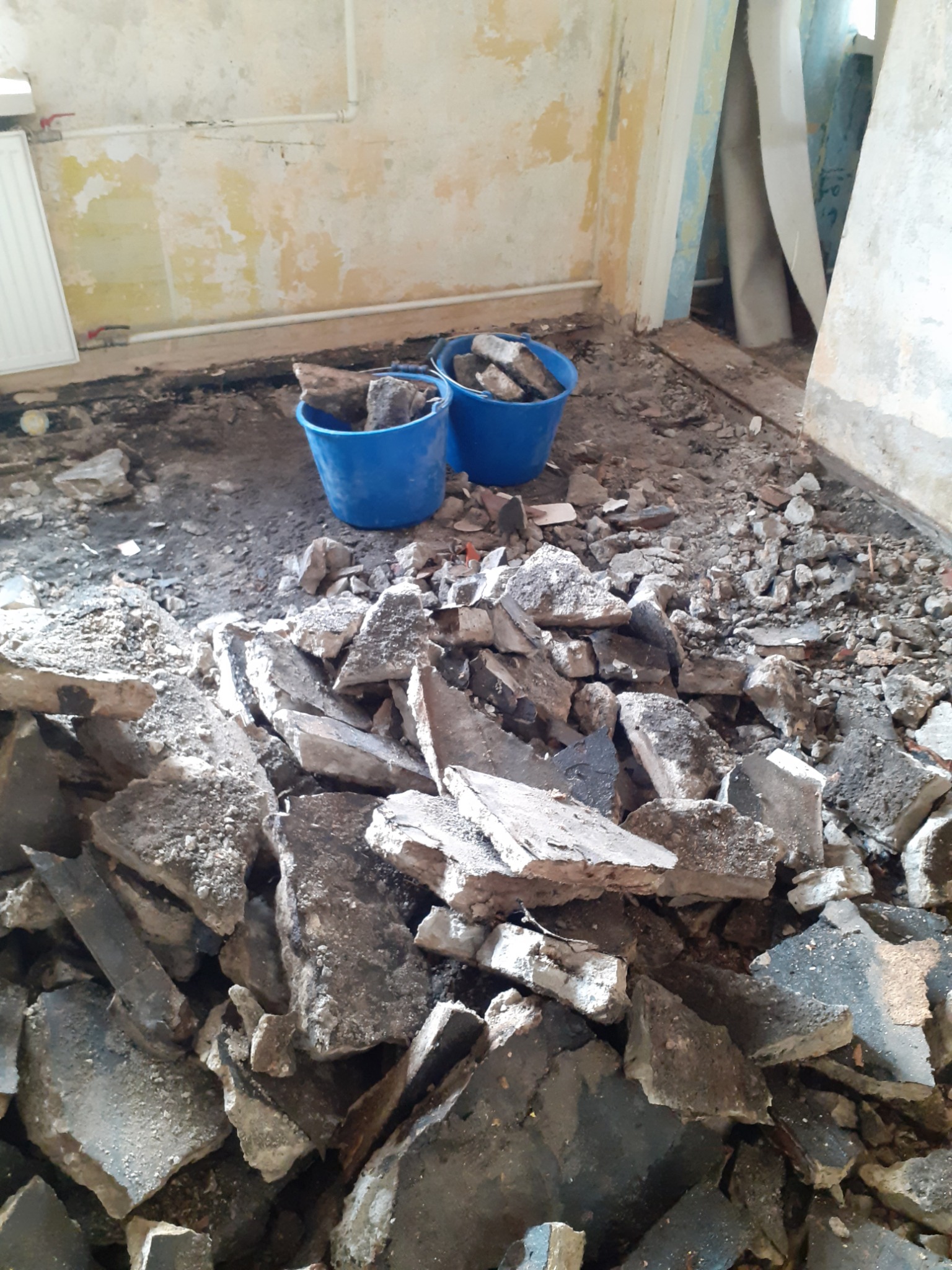 Stos gruzu i fragmentów betonu na podłodze w trakcie remontu, dwa niebieskie wiadra częściowo wypełnione gruzem w tle, ściany z widocznymi śladami starej farby.