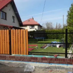 Montaż nowego ogrodzenia z drewnianymi panelami i metalowymi przęsłami na podmurówce z cegły klinkierowej w Piasecznie, widoczne narzędzia i materiały budowlane.