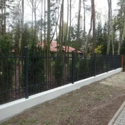 Nowoczesne, czarne ogrodzenie metalowe z betonową podmurówką na tle lasu i domu z czerwoną dachówką, widoczne z poziomu chodnika z kostki.
