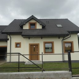 DECOTEL Mariusz Dudek - Jednorodzinny dom z beżową elewacją, brązowymi ramami okiennymi i ciemnobrązowym dachem z dachówki, widoczny z zewnątrz przez częściowo zamontowane ogrodzenie metalowe.