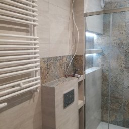 DECOTEL Mariusz Dudek - Wykończona łazienka z beżowymi płytkami, grzejnikiem drabinkowym, zabudowaną spłuczką WC i kabiną prysznicową z podświetlanymi półkami, widoczne kable wystające ze ściany.