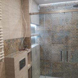 DECOTEL Mariusz Dudek - Nowoczesna łazienka z kabiną prysznicową z przesuwanymi drzwiami, płytki w odcieniach beżu i szarości z wzorem, podświetlone półki, instalacja elektryczna w trakcie podłączania.