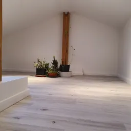 Jasne panele podłogowe w wykończonym pokoju na poddaszu w Piasecznie, z widocznymi listwami przypodłogowymi i drewnianym słupem konstrukcyjnym oraz roślinami doniczkowymi w tle.