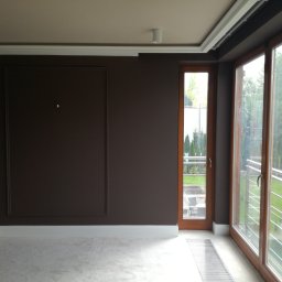 DECOTEL Mariusz Dudek - Pokój po malowaniu na ciemny brąz z drewnianą stolarką okienną i drzwiową, widoczne listwy przypodłogowe, dekoracyjne panele ścienne i grzejnik podłogowy.