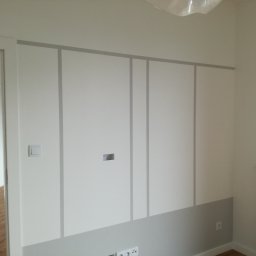 DECOTEL Mariusz Dudek - Świeżo pomalowana ściana z szarymi pionowymi i poziomymi pasami, dolna część ściany również szara, widoczny biały grzejnik i włącznik światła, w tle widok na parkiet przez otwarte drzwi.