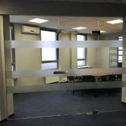 Przeszklone drzwi przesuwne z poziomymi, matowymi pasami w biurze, widok na salę konferencyjną z ciemną wykładziną i jasnym oświetleniem sufitowym.