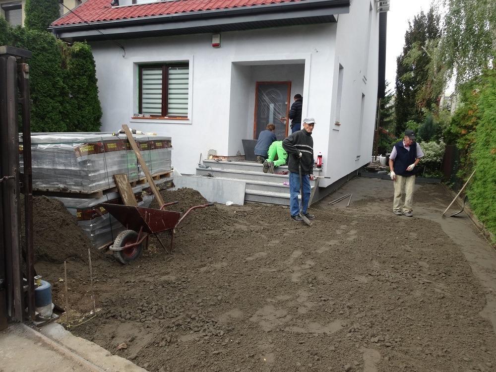 Prace ziemne przed domem jednorodzinnym: wyrównywanie terenu pod kostkę brukową, widoczni pracownicy z narzędziami, palety z kostką brukową, taczka i świeżo wyremontowane schody wejściowe.