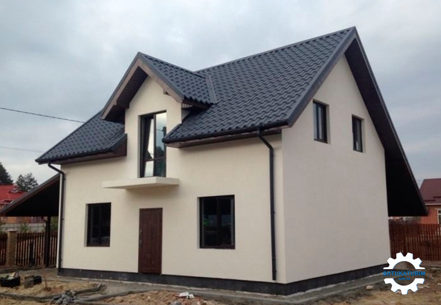 Otynkowany dom jednorodzinny z ciemną dachówką, widoczne okna, drzwi wejściowe i fragment ocieplenia fundamentów, na tle pochmurnego nieba.