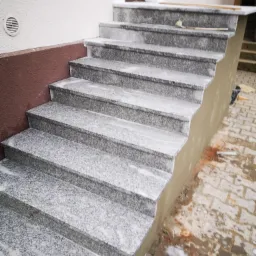Granitowe schody zewnętrzne w trakcie montażu, widoczne świeże zaprawy i niedokończone obrzeże, ceglana podłoga częściowo zabrudzona.