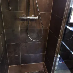 Nowoczesna kabina prysznicowa z ciemnymi, matowymi płytkami, chromowaną armaturą i odpływem liniowym w podłodze.