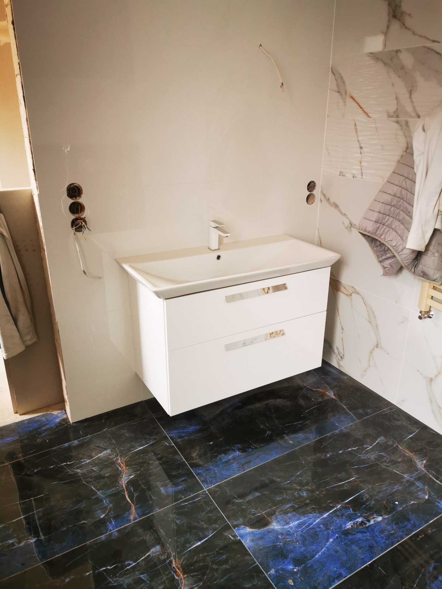 Biała wisząca szafka z umywalką i baterią w łazience z marmurowymi płytkami na ścianach i podłodze z ciemnego marmuru z niebieskimi żyłami, widoczne niedokończone instalacje elektryczne.