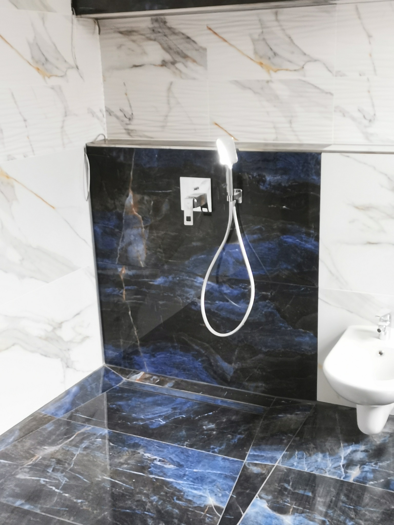 Nowoczesna łazienka z marmurowymi płytkami w odcieniach bieli i granatu, armatura chromowana, widoczny natrysk i umywalka.